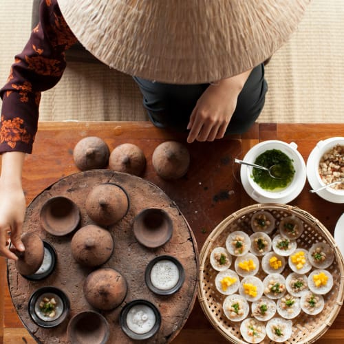A person wearing a large straw hat arranging traditional clay pots and dishes on a wooden table, with small bowls of food and herbs.