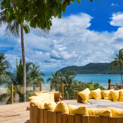 Outdoor lounge with a yellow cushioned circular sofa on a wooden deck overlooking the ocean and lush green islands under a partly cloudy sky.