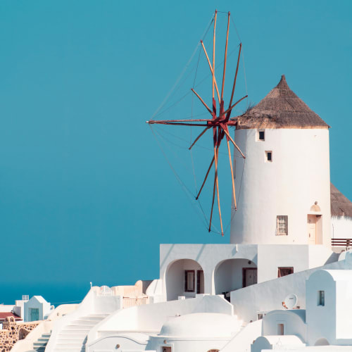 Traditional white buildings with a thatched-roof windmill under a clear blue sky in a coastal town.