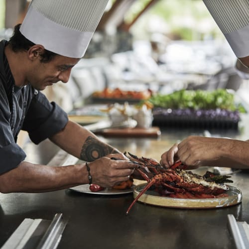 Two chefs with white hats delicately prepare lobster on a kitchen counter outdoors.
