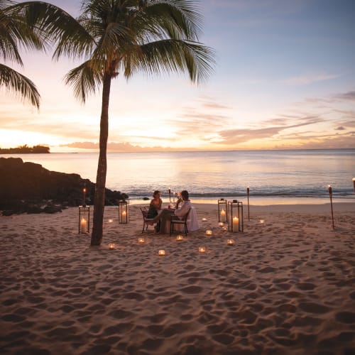 Couple sitting at a small table on a sandy beach during sunset, surrounded by lit candles and tiki torches with a palm tree nearby and calm ocean in the background.