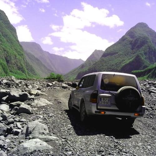 Ein weißes Geländefahrzeug fährt auf einem felsigen Weg in einem engen Tal, umgeben von steilen grünen Bergen unter einem teilweise bewölkten Himmel.