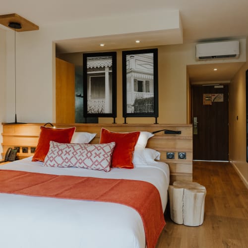 Modernes Hotelzimmer mit Doppelbett, orange-weißer Bettwäsche, hölzernem Kopfteil und zwei schwarz gerahmten Fenstern, die Außenarchitektur spiegeln.