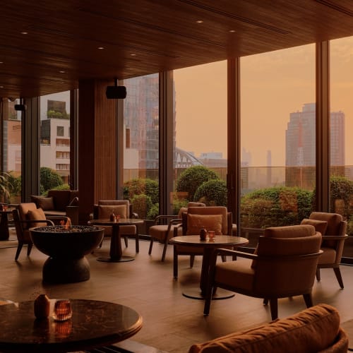 Luxurious lounge area in a high-rise building during sunset with large glass windows, leather armchairs, and a view of Bangkok cityscape.