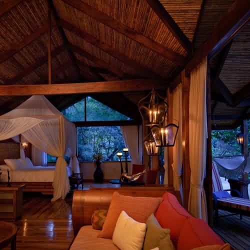 Interior of a rustic villa featuring a four-poster bed, wooden furniture, and cozy seating with cushions, overlooking a balcony with lush greenery.