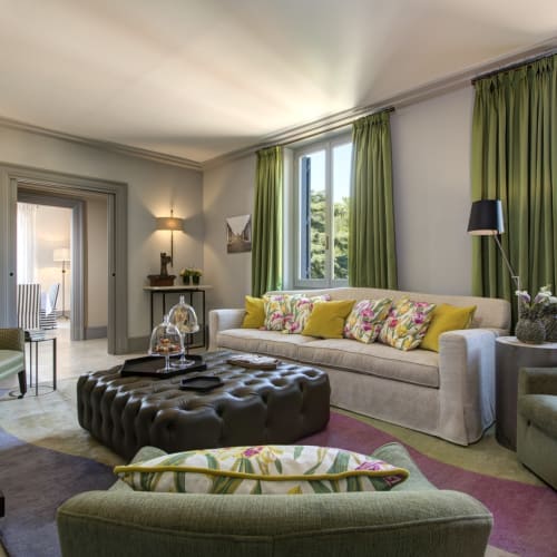 Elegantes Wohnzimmer mit grünen Vorhängen, bunten Kissen und antikem Mobiliar in einer hellen Hotelsuite.