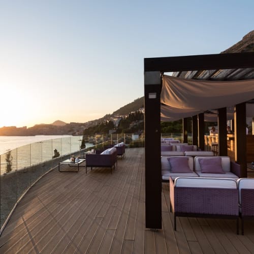 Eine luxuriöse Terrasse im Freien bei Sonnenuntergang, mit modernem Lounge-Mobiliar, Glasgeländer und Blick auf Hügel und Wasser.