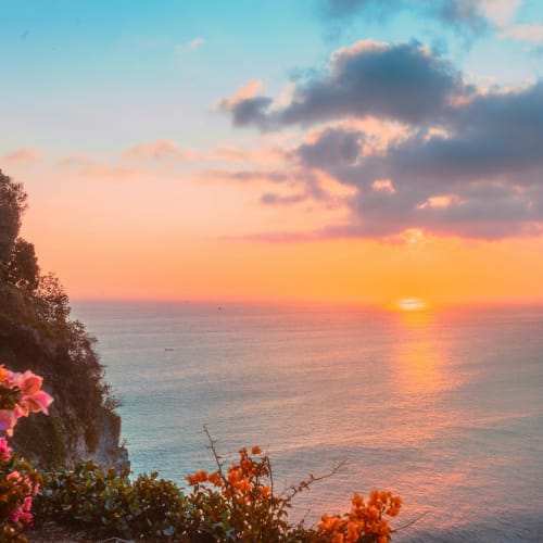Sunset over the ocean from a cliffside with colorful flowers and lush greenery in Bali.