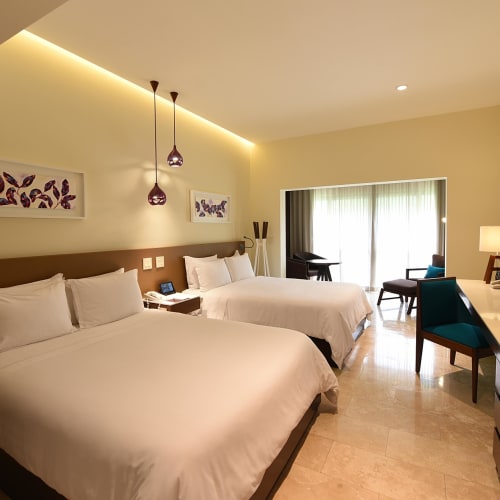 Modern hotel room with two large beds, a sitting area by the window, and flat-screen TV, illuminated with soft lighting.