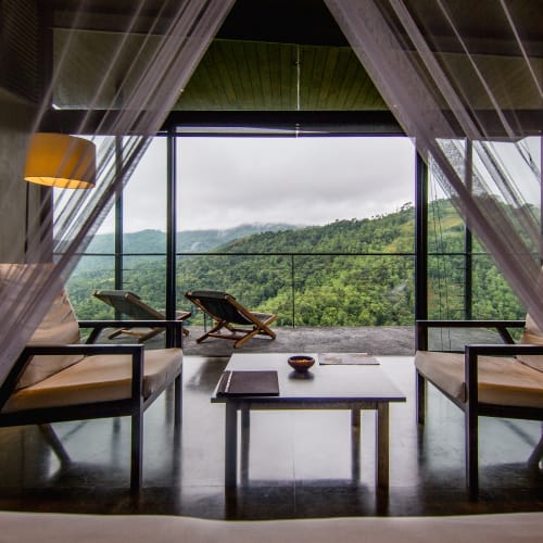 A cozy indoor seating area with two armchairs and a coffee table, overlooking a lush green mountain landscape through a large window with glass doors. Sheer curtains frame the view.