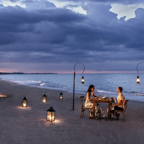 Ein Paar sitzt bei Dämmerung an einem kleinen Tisch am Strand, umgeben von Laternen, mit ruhigem Meer und bewölktem Himmel im Hintergrund.