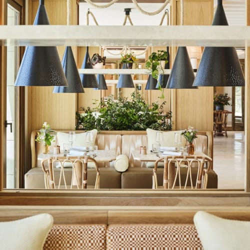 Interior of a luxurious restaurant at Four Seasons Resort Mallorca featuring a mirrored wall reflecting a beige sofa, wooden chairs, green plants, and multiple black pendant lights.