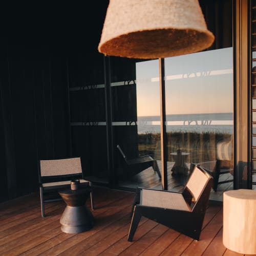 Moderner Außenbereich mit zwei gewebten Stühlen, einem kleinen Metall-Beistelltisch und einem großen hängenden Lampenschirm auf einem Holzboden vor reflektierenden Glasfenstern bei Sonnenuntergang.