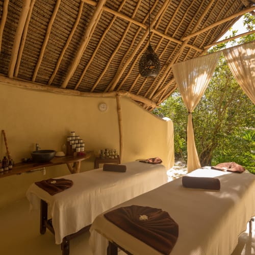 Ein ruhiger Spa-Behandlungsraum mit zwei Massagebetten, bedeckt mit weißen Laken, in einer offenen Struktur mit Strohdecke und umgeben von Grün.