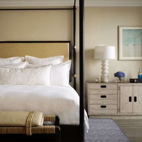 A spacious hotel bedroom with a four-poster bed, white bedding, and a matching white dresser with a table lamp, situated in a modern, well-lit room.