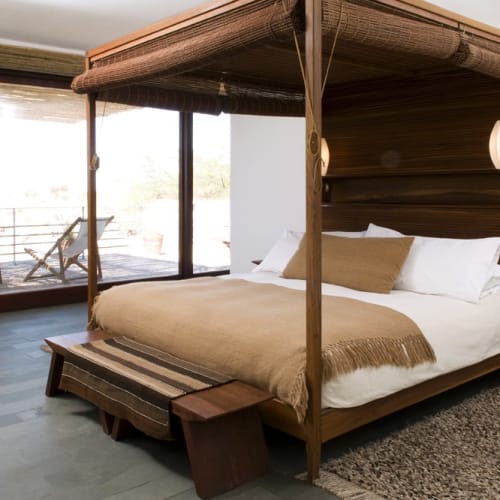 A luxurious bedroom with a four-poster bed in a modern hotel room, featuring large sliding glass doors leading to a balcony with outdoor seating.