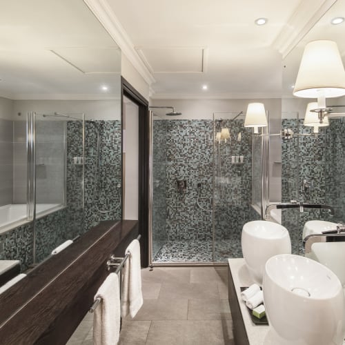 Modernes Badezimmer mit einem doppelten weißen Keramikwaschbecken, einer mit Glas umschlossenen Dusche mit Mosaikfliesen und einer in der Spiegelreflexion sichtbaren Badewanne, ausgestattet mit wandmontierten Lampen und braun gefliesten Boden.