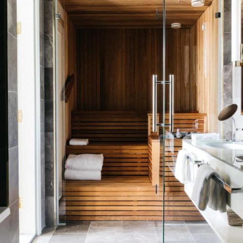 Modernes Badezimmer mit Glastür, die in einen hölzernen Saunaraum führt, mit gefalteten weißen Handtüchern und einem Doppelwaschbecken mit Handtüchern und Toilettenartikeln.