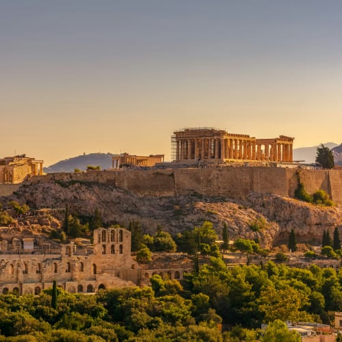 Blick auf die Akropolis von Athen bei Sonnenuntergang, mit dem Parthenon und umliegenden antiken Bauwerken auf einem felsigen Hügel und Grünflächen im Vordergrund.