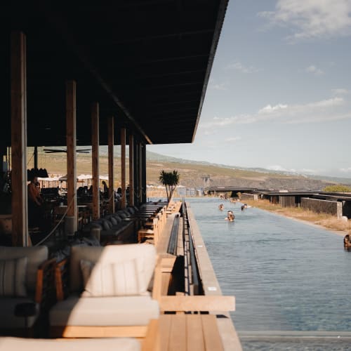Outdoor infinity pool with people swimming, adjacent to a modern covered seating area with cushioned chairs, overlooking a natural landscape and ocean under a partly cloudy sky.