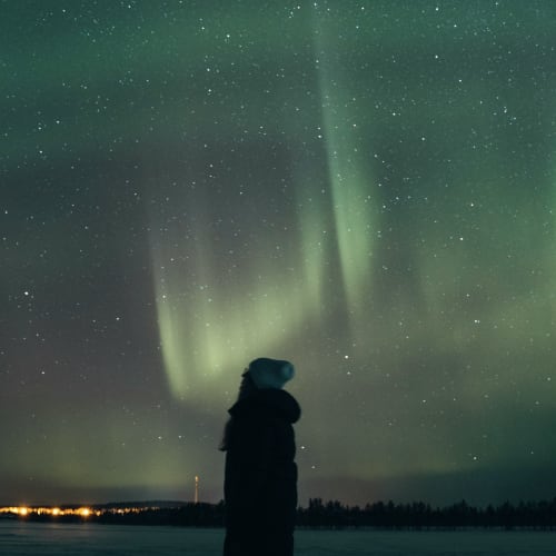 Eine Person steht im verschneiten Lappland unter einem sternenklaren Himmel, der von grünen Nordlichtern erleuchtet wird, mit entfernten Lichtern am Horizont.