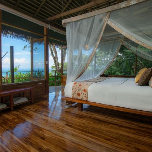 Luxuriöses Schlafzimmer in einem Bungalow mit Himmelbett und Blick auf Bäume und den Ozean durch große Fenster in einer tropischen Lodge.