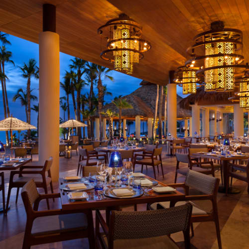 Elegantes Außenrestaurant mit Holzhimmel, hängenden Lampen und tropischen Palmen draußen bei Dämmerung.
