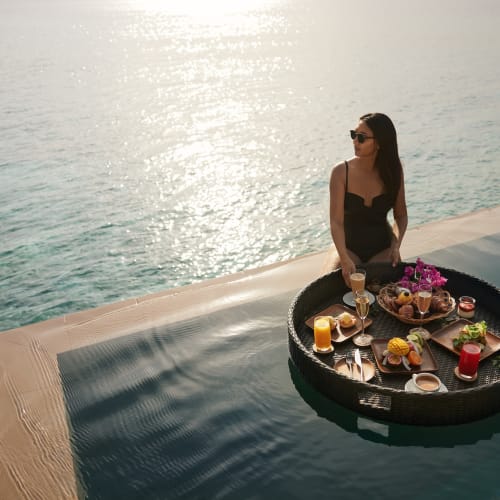 Eine Frau mit Sonnenbrille in einem schwarzen Kleid sitzt neben einem Infinity-Pool mit einem schwimmenden Frühstückstablett mit Blick auf den Ozean bei Sonnenuntergang.