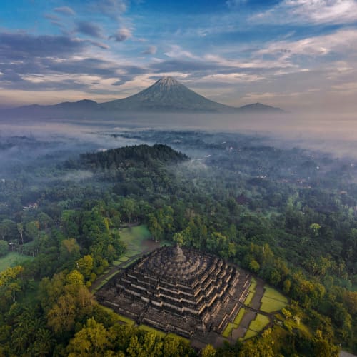 An aerial view of an ancient stepped temple surrounded by lush green forest with Mount Merapi in the background.