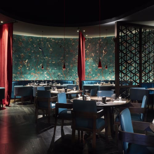 Elegant restaurant interior with round tables, blue chairs, brass pendant lights, and a mural wall with red curtains.