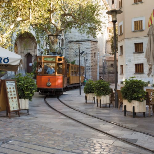 Eine orangefarbene Vintage-Straßenbahn fährt auf Gleisen durch eine gepflasterte Straße mit Außensitzplätzen und Pflanzen, im Hintergrund Steinbauten und Bäume an einem sonnigen Tag.