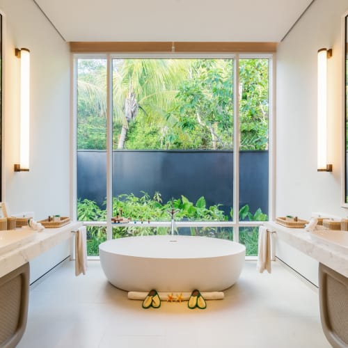 Modern white bathroom with a freestanding oval bathtub centered between two sinks and large windows overlooking lush green tropical plants outside.