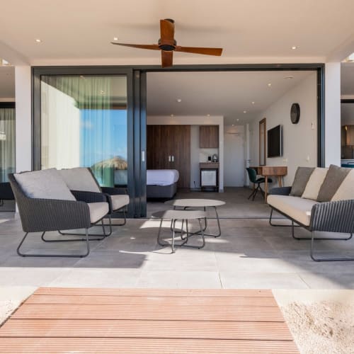 Modern outdoor lounge area with seating, overlooking a hotel room with sliding glass doors and a bedroom in the background.