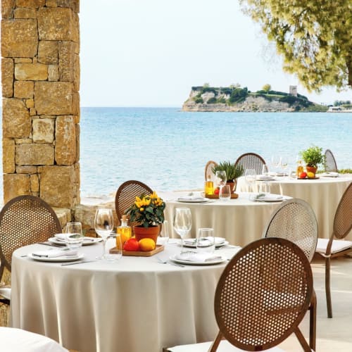 Elegant outdoor restaurant setup on a beach with white tablecloths and chairs, overlooking the sea and a distant island.