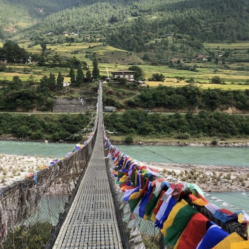 A suspension bridge decorated with colorful prayer flags stretching across a river with lush green hills in the background.