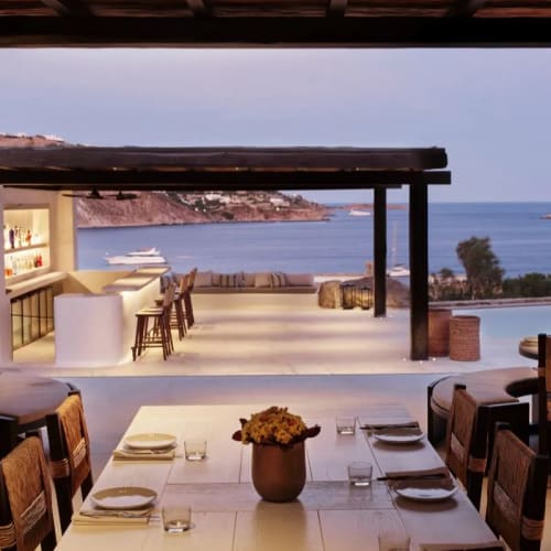 Luxuriöser Außenbereich des Kalesma Restaurants mit Blick auf die Küste, einer Bar, Hockern und einem für sechs Personen gedeckten Tisch mit Tellern, Gläsern und Besteck im Abendlicht.