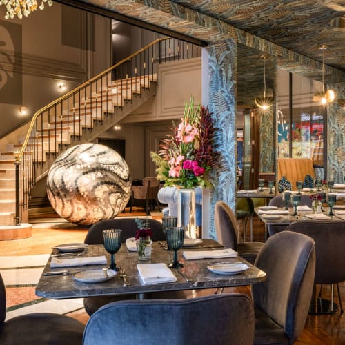 Elegantes Restaurantinnere mit sorgfältig eingedeckten Tischen, einer großen Blumenarrangement und einer dekorativen Marmorkugel in der Nähe einer Treppe.