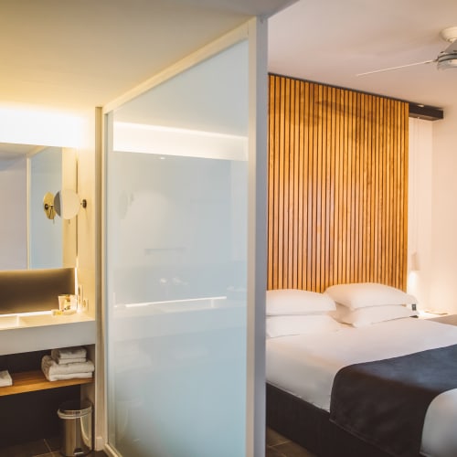 Modernes Hotelzimmer mit großem Bett, weißer Bettwäsche, Holz-Kopfteil und Badezimmer hinter mattiertem Glas.