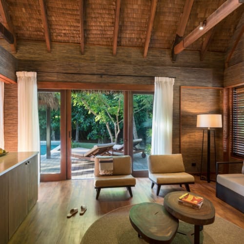 Living room with large glass door opening to an outdoor pool area surrounded by lush greenery, featuring wooden walls, a lounge chair outside, and comfortable indoor furniture.