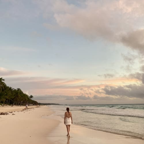 A person in a white dress walking barefoot along a sandy beach near the ocean with palm trees on the left under a pastel-colored sky at sunset or sunrise.