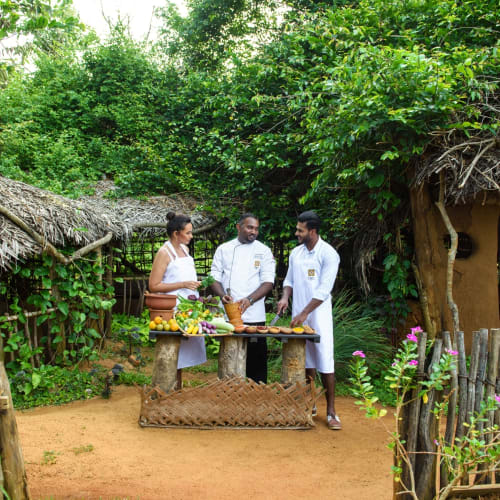 Drei Menschen in traditioneller Kleidung stehen in einer lushen, grünen Umgebung um einen Tisch mit Trommeln und anderen Gegenständen, wohl in einem afrikanischen Dorf.