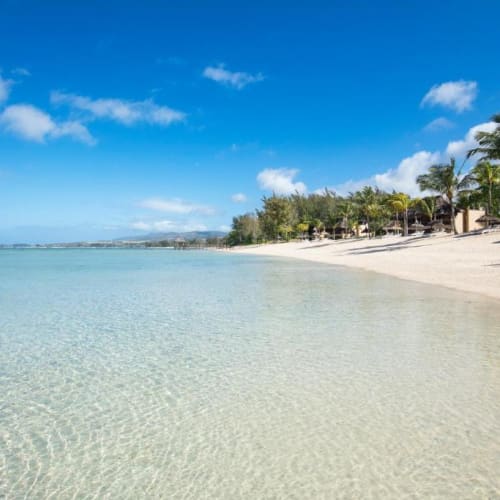 A tranquil beach with clear turquoise waters, white sandy shore, and palm trees under a blue sky with some clouds.