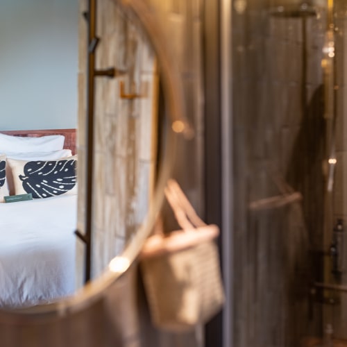 Spiegelbild eines ordentlich gemachten Betts mit weißen Laken und zwei Kissen mit Monstera-Blatt-Muster in einem runden Wandspiegel, daneben eine verschwommene gewebte Tasche, in einem gemütlichen Schlafzimmer.