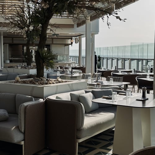 Moderner Außenbereich eines Restaurants mit gepolsterten Sofas und Tischen unter einem Holzpergola, mit Topfbäumen und einer Glasbarriere mit Blick auf das Meer.