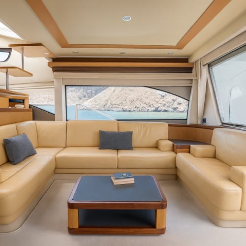 Spacious and modern yacht interior with beige leather sofas arranged in a U shape, a wooden and black coffee table with books on it, and large windows showing a mountainous water view outside.