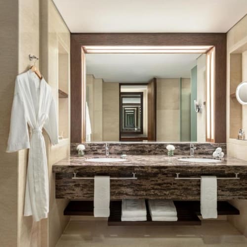 A luxurious bathroom features a large mirror, a marble countertop with two sinks, and a robe hanging on the wall. Towels are neatly placed under the countertop, and there's a modern shower area on the side.