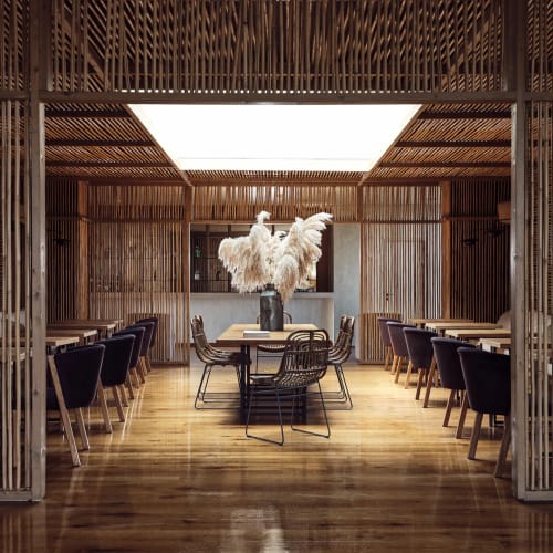 Interior view of a dining area with wooden lattice walls and ceiling, featuring a central table decorated with a large vase of pampas grass and surrounded by multiple dark cushioned chairs.