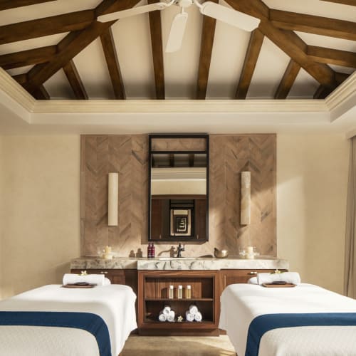 Spa-Zimmer mit zwei Massageliegen, einem Spiegel und Blick auf üppiges Grün draußen durch ein großes Fenster.