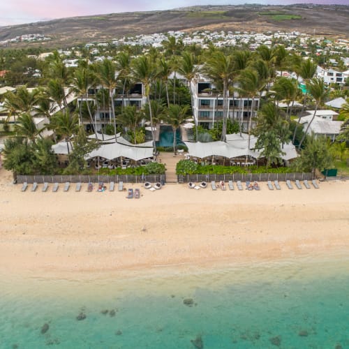 Luftaufnahme eines Strandresorts mit Palmen, Liegestühlen am Sandstrand, klarem türkisfarbenem Wasser und einem Wohngebiet am Hügel im Hintergrund.