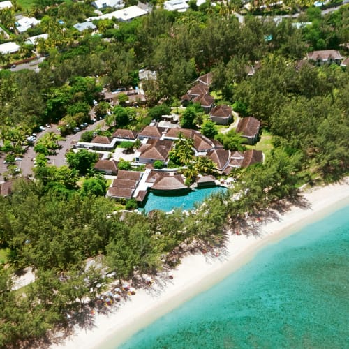 Aerial view of a coastal resort surrounded by dense green trees with several thatched-roof buildings and a turquoise swimming pool near a white sandy beach and clear blue ocean.